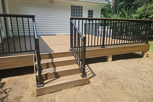 Ground-level brown composite deck with black metal railing panels and broad access stairs leading into the yard.
