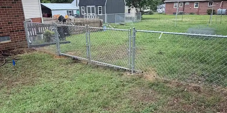 Silver metal chain-link fence with an integrated gate running across a green lawn next to a red brick home.