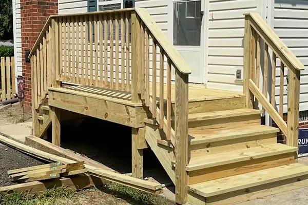 Newly constructed wooden porch deck and entry stairs with wooden railings leading up to the doorway of a house with white siding.