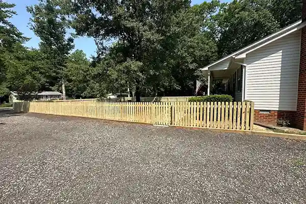 Long wooden picket fence running along a gravel driveway in front of a residential house surrounded by shade trees.