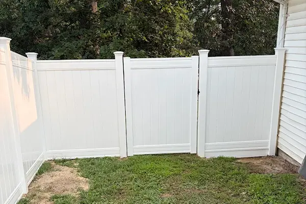 White vinyl privacy fence enclosure featuring a central swing gate on a green lawn next to a residential house.