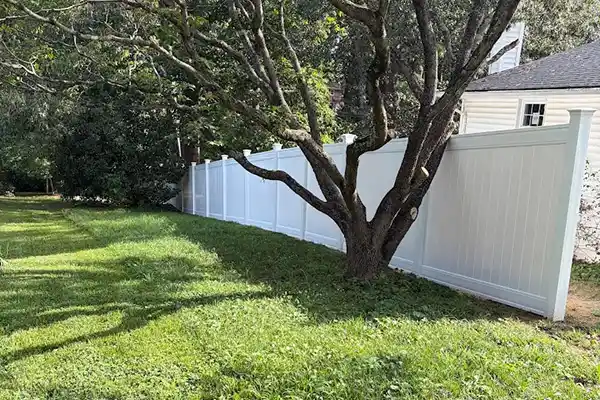 Long line of white vinyl privacy fencing running along a shaded lawn behind a large mature tree.
