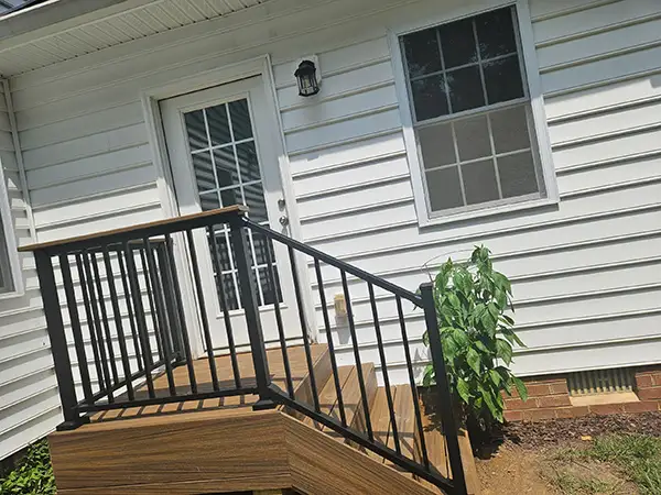 Brown composite deck landing and steps with black metal railings leading out from the back door of a white-sided house.